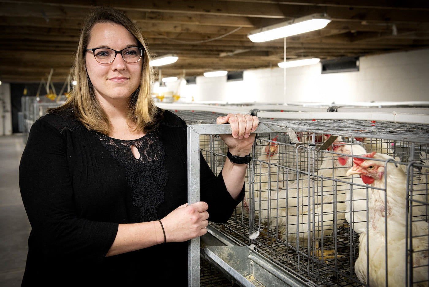 A woman, Sara Orlowski, in a black blouse with glasses stands next to chicken cages in an indoor agricultural facility, with several white chickens in the background.