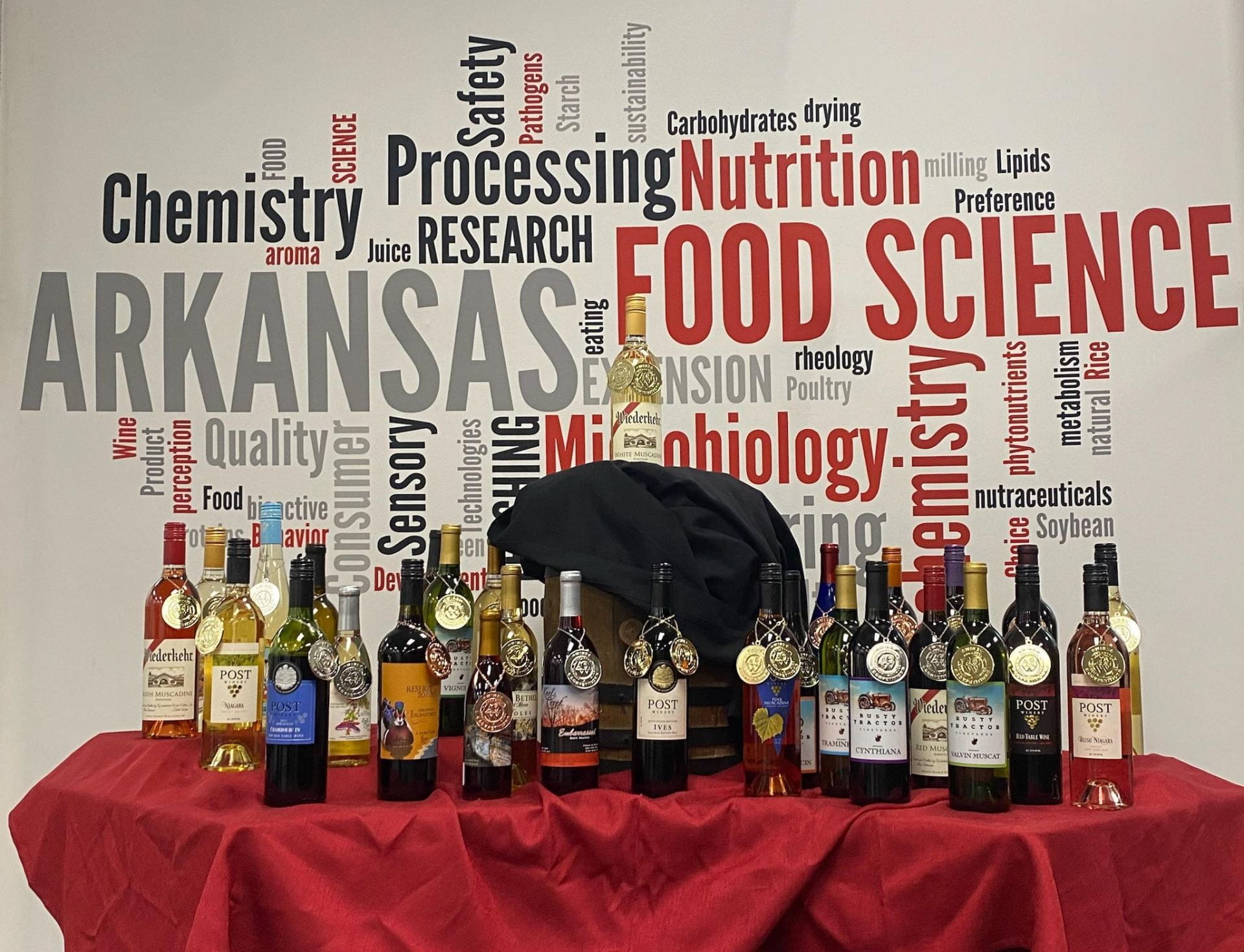 Photo of about 30 wine bottles with gold and silver medals placed on a red tablecloth in front of a word cloud poster with food science terms on it.