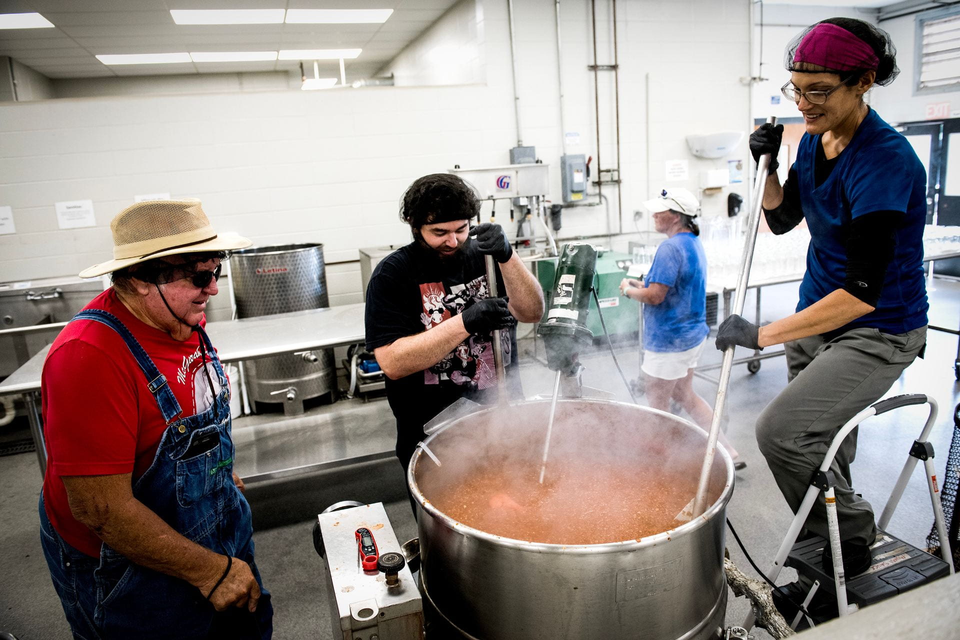 Three people stand around a large steaming kettle containing a tomato-based food.