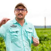 Man gestures with his hand while presenting during a field day with green crops in the background.