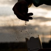 A person's hands silhouetted against a sunset, with one hand scattering fine soil particles and the other holding a box