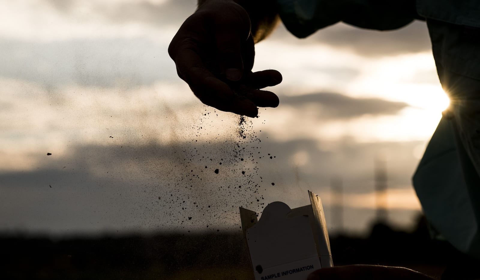 A person's hands silhouetted against a sunset, with one hand scattering fine soil particles and the other holding a box
