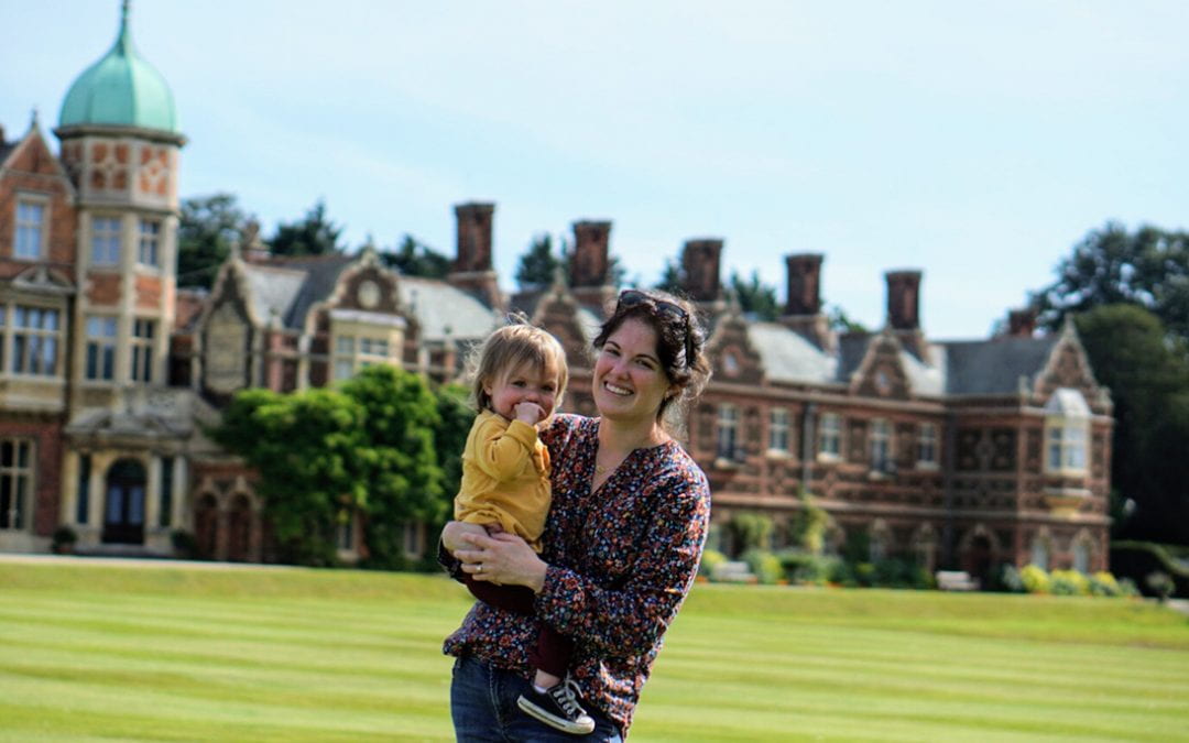 Kelsey wearing a black floral shirt and holding a toddler in a yellow shirt in front of a brick estate.