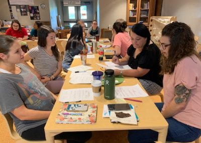 Four women sit at a table in the foreground, reading their papers, while other women sit at tables in the background during a workshop at the September Professional Educator Development Day at the Jean Tyson Child Development Study Center at the University of Arkansas.