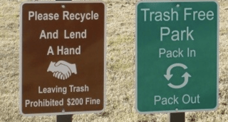 A brown sign (left) reads: "Please Recycle and Lend a Hand; Leaving Trash Prohibited $200 Fine." A green sign (right) reads: "Trash Free Park; Pack In, Pack Out."