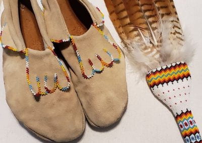 Two items of PowPow regalia laid out, including a tanned pair of closed toed shoes with colorful fringing along the top edges and a feather fan colorful and intricate beading along the handle.