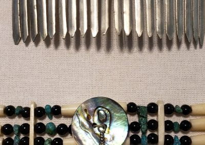 Two PowPow accessories laid out, including a decorative metal hair comb with many teeth to hold it in place and a beaded bracelet. The comb in silver with a blue stone in the center and the bracelet has blue, black and white beading with an iridescent circular shell piece in the middle with a small metal feather attachment..
