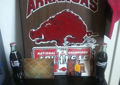 Display of personal U of A objects, including a wooden plaque featuring "Arkansas Razorbacks" with a hog illustration in the middle and with a UA hat hanging from it, a red and white 1994 UA National Champions commemorative license plate, two clear glass Coca-Cola UA commemorative bottles, and two photo prints featuring a brown floor tile with "Delta Upsilon Arkansas Chapter" on it and other surrounding named tiles and another photo of a person in a yellow sweater holding a commemorative plaque.